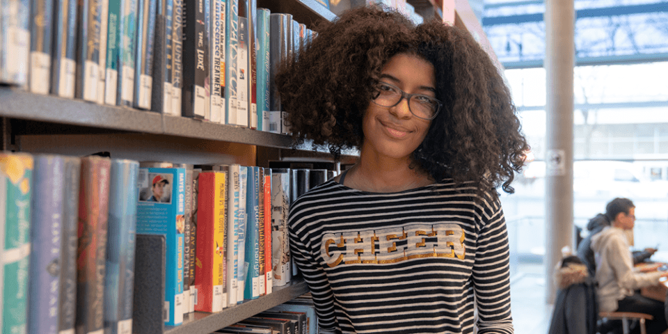 Photo of a teenager with glasses wearing a striped shirt that reads Cheer, standing next to a row of library bookshelves. 