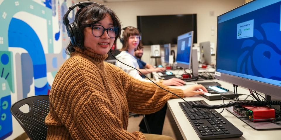 A teen with headphones and a mustard-hued sweater sits at a computer while smiling at the camera. 