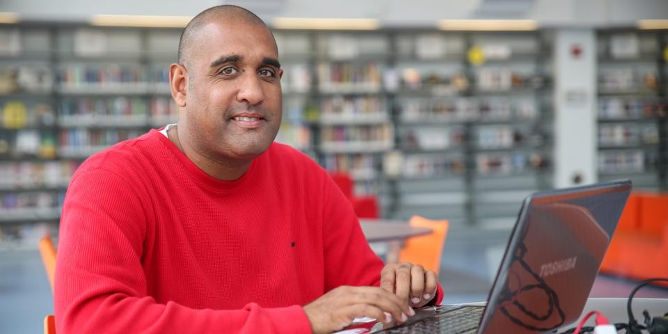 A man in a red shirt smiles at the camera while typing on a laptop.