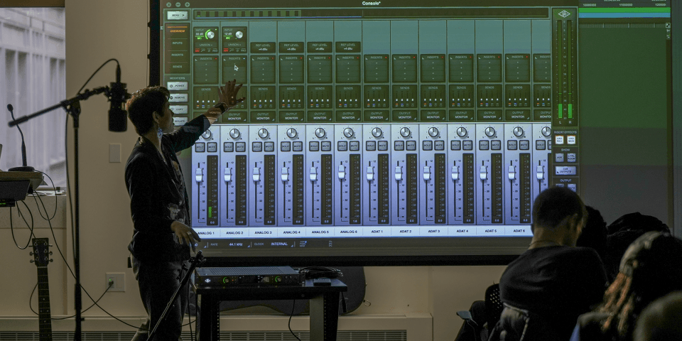 Man pointing to a digital sound mixer in a room with microphones and musical instruments.