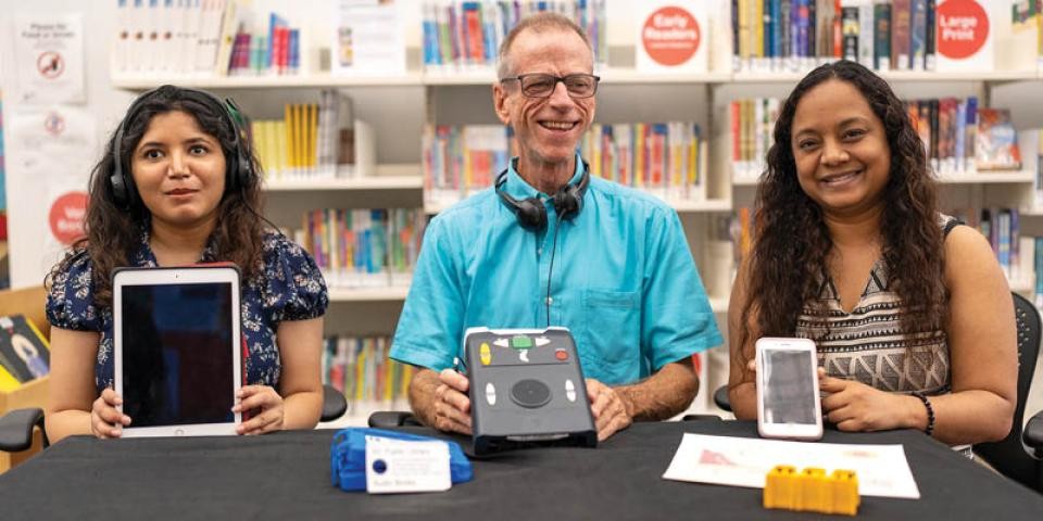Three people sit at a table in a library, holding various accessible devices for books. 