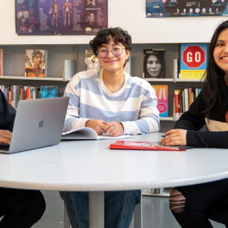 Three teens sit gathered around a circular table in a room filled with books; they all smile at the camera and one has a laptop. 