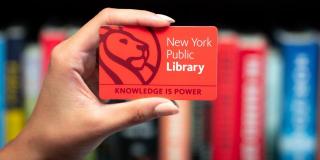 Photograph of a hand holding a red NYPL card that reads: New York Public Library, Knowledge Is Power