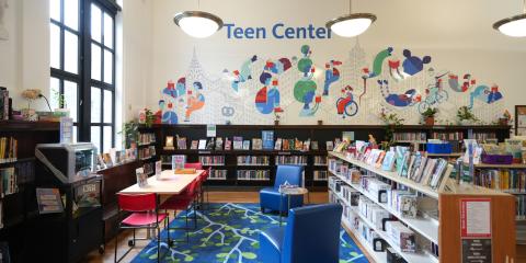 Harlem Library Teen Center, featuring a work table, blue lounge chair, shelves of books lining the space, and a colorful wall mural of people reading in a cityscape.
