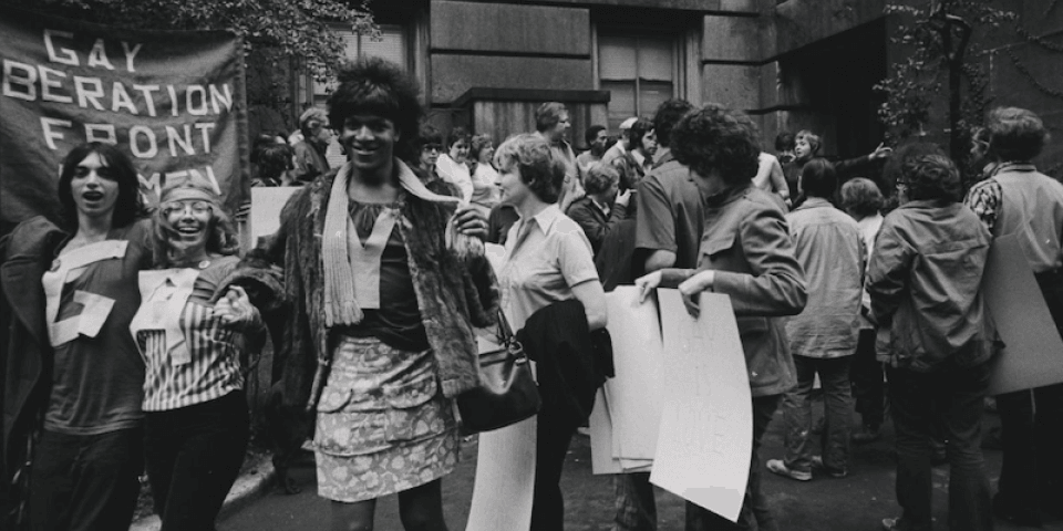 Historic black-and-white photo featuring a group of people, including Marsha P. Johnson, rallying in support of LGBTQ rights; in the background, a handmade sign reads: Gay Liberation Front. 