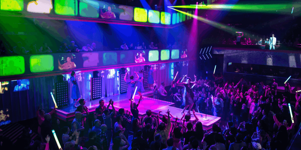Green and purple lights and videos shine on a catwalk stage as people dance on the floor.