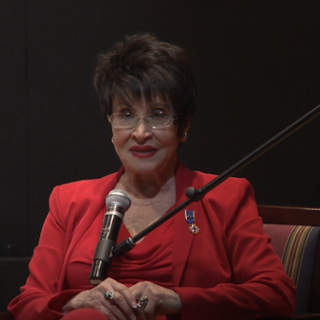 A woman with short black hair and a red suit speaks in front of a mic